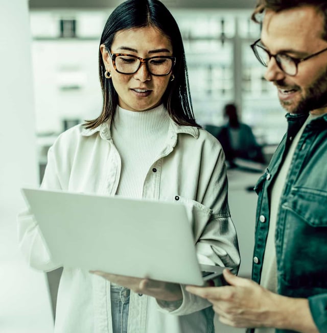 A man and a woman stand next to each other, looking at a laptop she is holding.