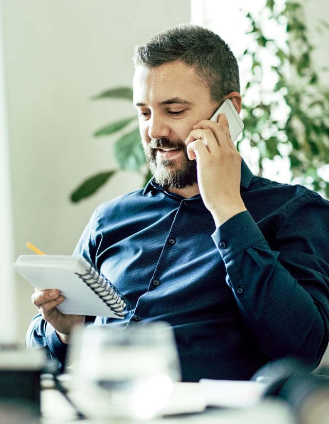 A man talking on a smartphone looks at a spiral-bound notebook.