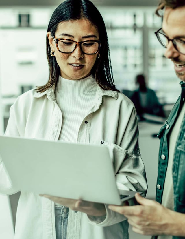 A man and woman stand next to each other reviewing information on a laptop she's holding.