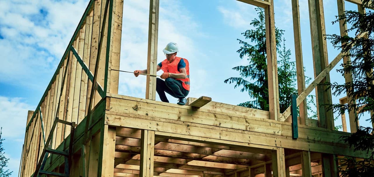 A construction worker takes a measurement in a partially-framed building.