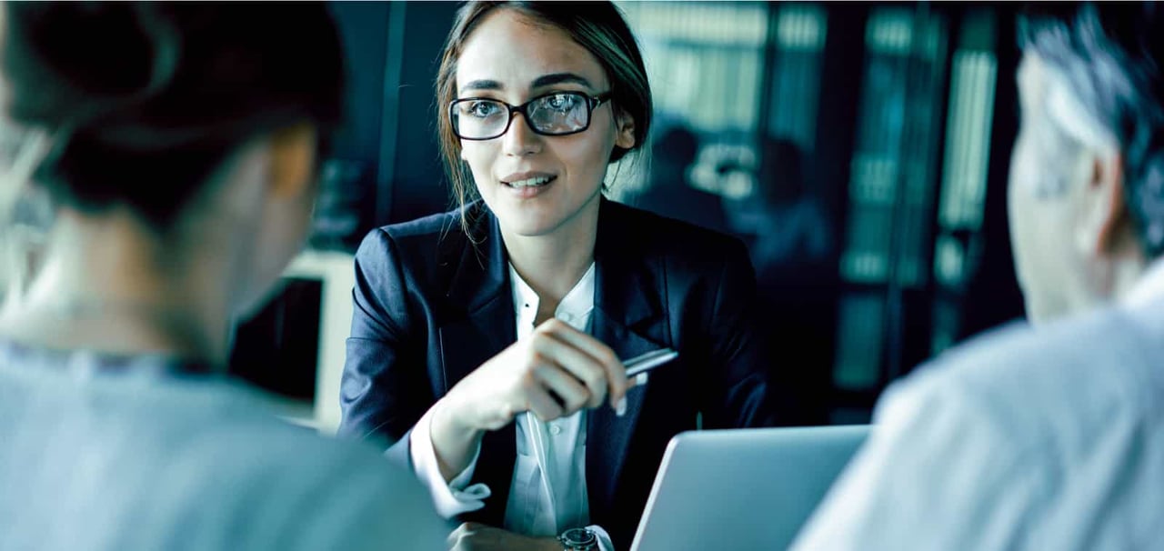 A young woman in a blazer talks to two people sitting in front of her desk.