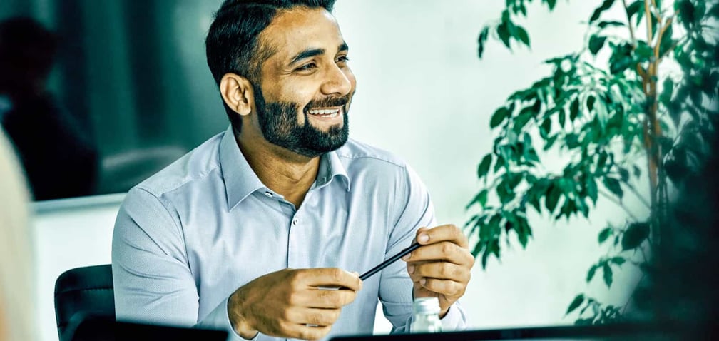 A smiling man in a blue shirt holds a pencil during a meeting in an office.