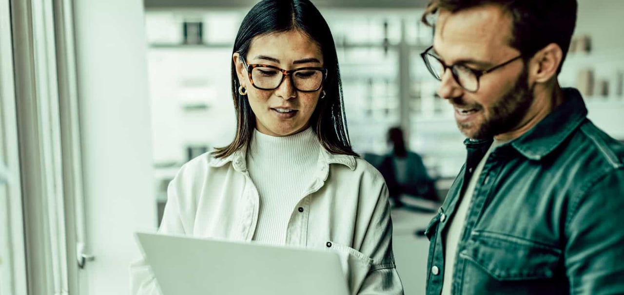 A man and woman stand next to each other reviewing information on a laptop she's holding.