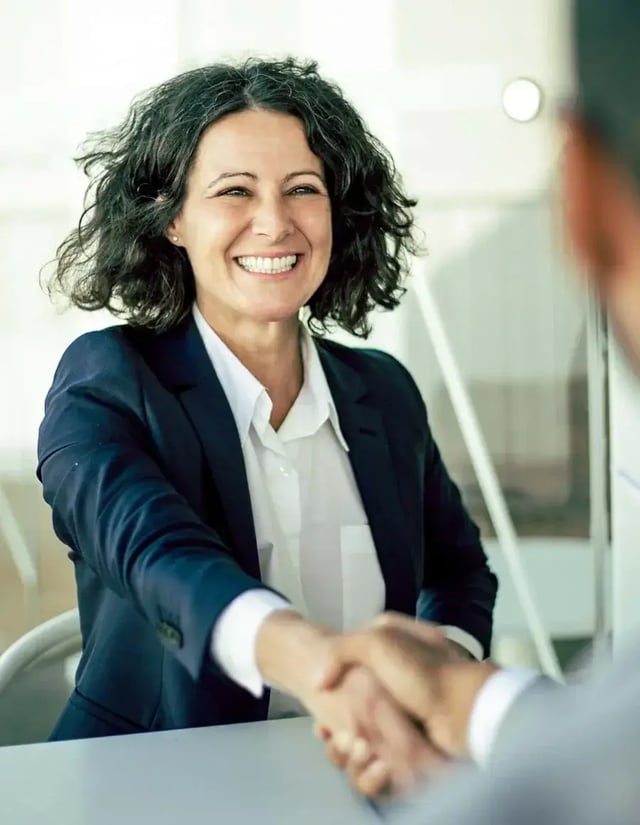 A smiling woman in a blazer shakes hands with someone off-camera.