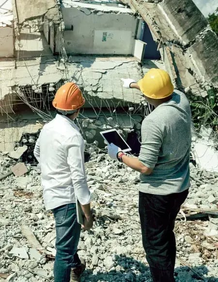 Two people in hard hats look at the rubble of a collapsed building