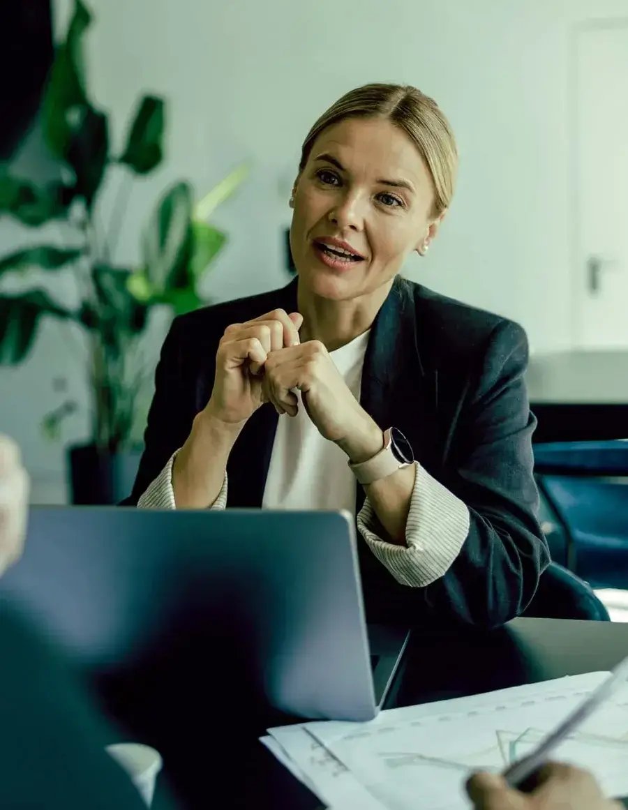 A woman in a blazer talks to other people at a conference table with laptops and papers.