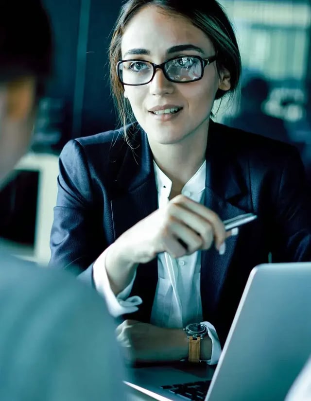 A young woman in a blazer talks to two people sitting opposite her desk.