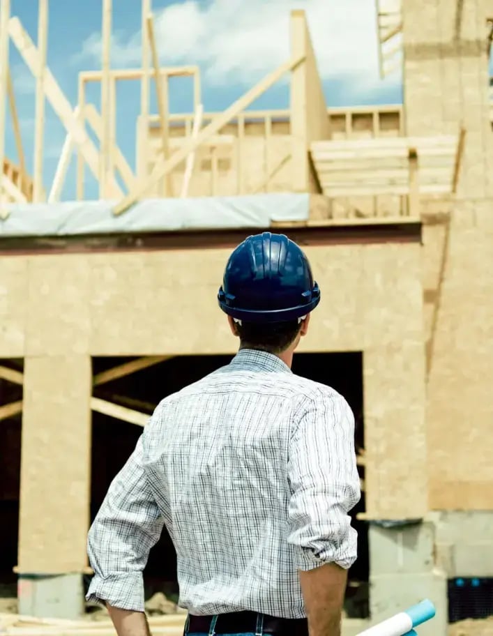 A man in a button-down shirt and a hard hat looks at an under-construction home