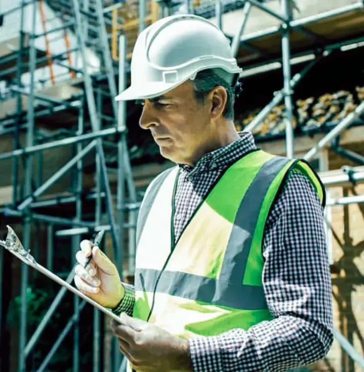 An inspector in a vest and hard hat reviews information on a clipboard at a project site